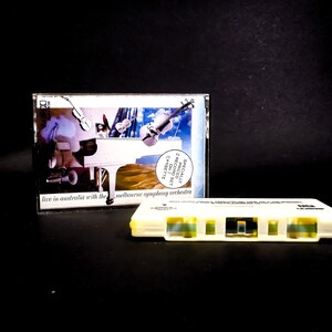 May include: A white cassette tape with a yellow label and a clear plastic case. The case has a collage of images including a piano, a violin, and a landscape. The text on the case reads "Specially Priced 2 Record/1 Cassette Set Live in Australia with the Melbourne Symphony Orchestra".