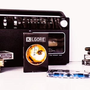 May include: A black cassette player with a cassette tape labeled "Kilgore" inside. There are several other cassette tapes stacked next to the player, including one labeled "Spinal Tap". There is also a stack of cassette tapes with a small orange figurine on top.