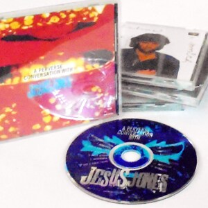May include: A CD case with a red, yellow, and blue design. The text on the case reads "A Perverse Conversation with Jesus Jones".
