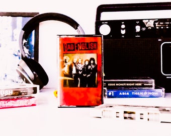 May include: A collection of cassette tapes, including Bad English, Eddie Money, Asia, and Rush. The tapes are arranged on a white surface next to a black cassette player and a pair of black headphones.