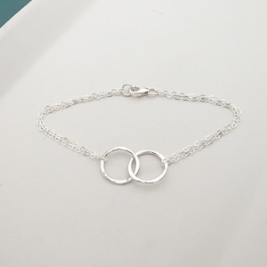 May include: A delicate silver bracelet featuring two interlocking circles, connected by a fine chain. The bracelet has a lobster clasp closure. The design is simple and elegant, suitable for everyday wear.