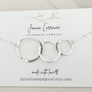 May include: A silver necklace featuring three interconnected hammered rings. The necklace is displayed on a white card with the text "Janine Lorraine Handmade Jewelry" and "made with love". The Etsy shop URL is also visible.