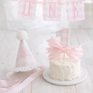 May include: A festive first birthday party scene. Includes a small white cake with a pink bow and a number one cake topper, a pink and white gingham party hat, and a banner that spells "ONE". All items are in shades of pink and white.