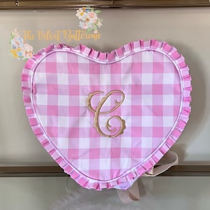 Pode incluir: Mochila em forma de coração com padrão xadrez rosa e branco. A mochila tem uma borda com babados e uma letra "C" dourada bordada no centro. As alças são de cor bege claro.