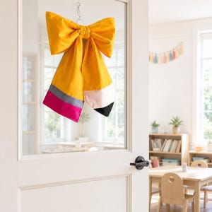May include: A large fabric bow shaped like a pencil, featuring a bright yellow top, a grey band, a pink bottom section, and a light pink tip with a black point. The bow is suspended by a clear suction cup hook on a glass pane of a white door.