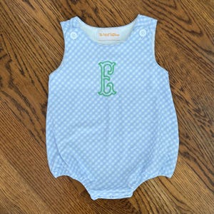 May include: A sleeveless light blue and white gingham bubble romper featuring a round neckline with two white buttons at the shoulders and a green embroidered letter E in the center. The interior label displays the text The Velvet Buttercup.