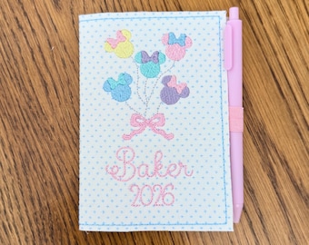 Könnte beinhalten: Ein hellblaues Notizbuch mit einem Tupfenmuster und einem rosa Stift. Der Notizbuchumschlag zeigt gestickte Mickey Mouse-Kopfballons in verschiedenen Farben, eine rosa Schleife und den Namen "Baker" mit dem Jahr "2026".