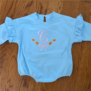 May include: Light blue baby romper with long sleeves and ruffled shoulders. The romper features a pink embroidered initial, a garland of orange pumpkins, and a pink bow. The romper is made of a soft material.