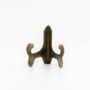 Op de afbeelding: Een kleine, goudkleurige metalen displaystandaard met een puntige bovenkant en drie poten. De standaard is ontworpen om kleine objecten te houden, zoals beeldjes of sieraden.