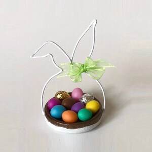 May include: White wire bunny shaped basket with a green bow and a variety of colourful chocolate eggs inside.