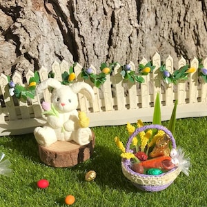 May include: A miniature Easter scene with a white bunny figurine holding a pink tulip and a yellow chick, sat on a wooden disc. A small basket filled with treats is nearby, with a white picket fence and decorative garland in the background.
