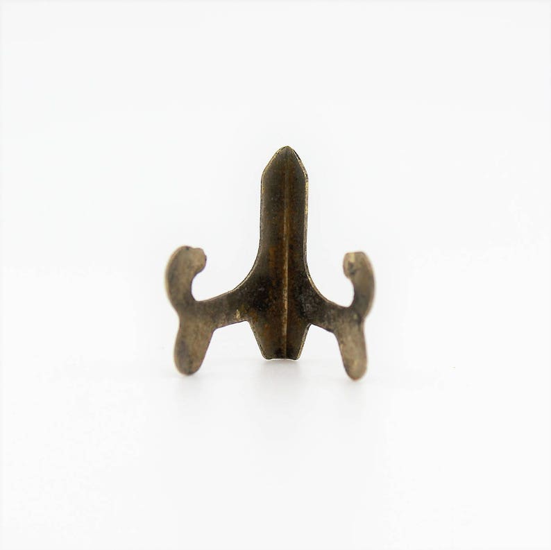 Op de afbeelding: Een kleine, goudkleurige metalen displaystandaard met een puntige bovenkant en drie poten. De standaard is ontworpen om kleine objecten te houden, zoals beeldjes of sieraden.
