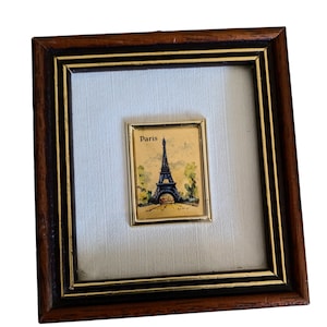 May include: A framed miniature painting of the Eiffel Tower in Paris, France. The painting is on a gold background with a black frame.