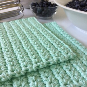 May include: Two mint green crocheted dishcloths stacked on top of each other. The dishcloths are made with a textured stitch.