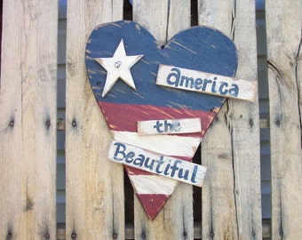 Peut inclure: Une pancarte en bois en forme de cœur peinte aux couleurs du drapeau américain, rouge, blanc et bleu. La pancarte porte les mots "America the Beautiful".