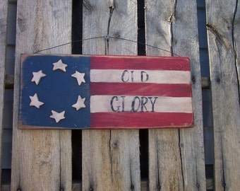 Peut inclure: Une pancarte en bois avec un motif de drapeau américain peint. La pancarte a 13 étoiles blanches en cercle sur fond bleu, et des rayures rouges et blanches. Les mots "Old Glory" sont peints en noir sur la pancarte.