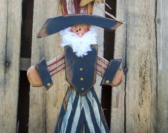 Peut inclure: Figurines en bois d'Oncle Sam, peintes en rouge, blanc et bleu, portant un grand chapeau avec une étoile au sommet. Il a une barbe blanche et se tient sur une surface rouge.