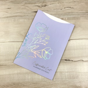 May include: A lavender planner sticker envelope with a floral design in iridescent foil. The text "Caffeinated Cat PLANNER STICKERS" is printed in silver. The envelope has a curved top.