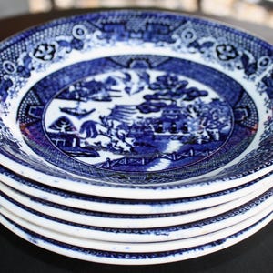 May include: A stack of six blue and white ceramic plates. Each plate features a detailed landscape scene in the center, surrounded by a decorative border. The plates are stacked neatly, showcasing the classic Willow pattern.