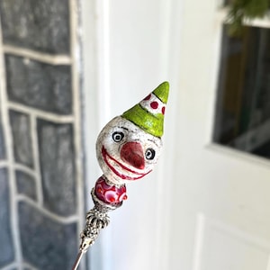 May include: A whimsical clown-faced bottle stopper. The clown has a white face with black eyes, a red nose and a red smiling mouth. It wears a green hat with red polka dots. The stopper has a red and white bead and a silver-colored metal base.