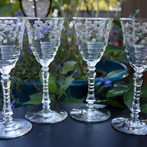 May include: Four clear wine glasses with etched floral designs. The glasses have a flared bowl, a faceted stem, and a round base. The etched design features small, round shapes and delicate lines. The glasses are arranged on a dark surface.