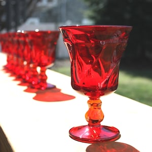 May include: A row of vibrant red glass goblets with a faceted, textured bowl and a decorative, multi-tiered stem. The translucent red glass catches the light, creating a distinct shadow on the surface below. Each goblet features a wide rim.