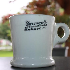May include: A white ceramic mug with a rounded base and a handle. The mug features the text "Cornwall Consolidated School" in black script. The mug is set against a blurred outdoor background.