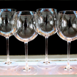 May include: Four clear wine glasses with long stems and rounded bowls are arranged in a row. The glasses are empty and reflect the surrounding environment. The background is dark, creating a contrast with the transparent glassware.