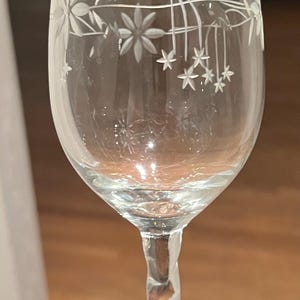 May include: A clear wine glass with a floral etched design. The glass features a twisted stem and a delicate pattern of flowers and leaves around the rim. The glass is approximately 7 inches tall.