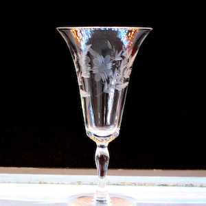 May include: A clear, etched glass with a flared top and a delicate stem. The glass features a floral design, with etched flowers and leaves. The glass is approximately 7 inches tall and is suitable for serving beverages.