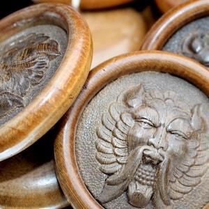 May include: Close-up of several wooden coasters with carved relief designs. The coasters have a circular shape with a raised rim and a detailed carving of a face with wings. The wood has a warm, natural tone.
