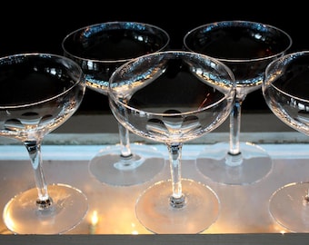 May include: A collection of six clear, stemmed champagne glasses. The glasses have a shallow, rounded bowl and a delicate stem, resting on a circular base. The glasses are arranged in a row, reflecting light and creating a sophisticated aesthetic.