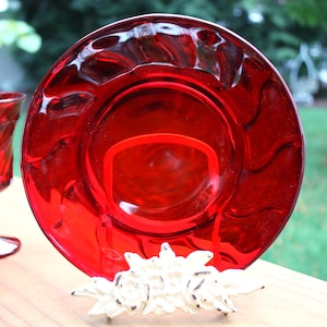 May include: A circular red glass plate with a wavy, textured rim, displayed upright on a white floral-shaped metal stand. To the left, a matching red glass goblet with a textured surface stands on a stem, both featuring a deep ruby hue.