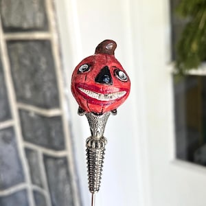May include: A Halloween-themed decorative pin featuring a smiling jack-o'-lantern face. The pumpkin has a red painted face with a black nose and mouth, white teeth, and a brown stem. The pin has a silver-colored, ornate base and a long, slender needle.