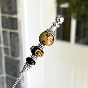 May include: A decorative silver-toned hat pin with a beaded design. The pin features a silver ball at the top, a speckled brown bead, a black and gold bead, and a black bead. The beads are separated by silver accents.