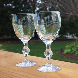 May include: Two clear crystal wine glasses with diamond-shaped stems sit on a wooden surface. The glasses reflect the surrounding environment, including trees and a house. The glasses are empty and ready for use.