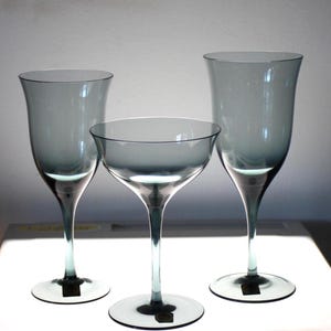 May include: Three gray-tinted glass stemware pieces. The set includes two tall, tulip-shaped glasses and a coupe glass. Each glass has a slender stem and a circular base. The glasses are arranged on a white surface, with a soft, diffused background.