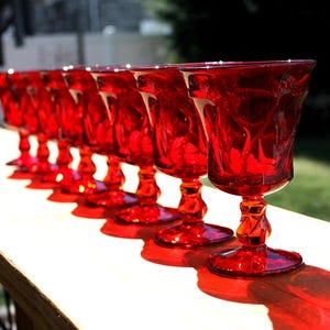 May include: A row of eight vibrant red glass goblets arranged in a line. Each goblet features a textured, faceted bowl with a distinct pattern and a short, twisted stem leading to a circular base, reflecting bright sunlight and casting shadows.