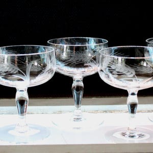 May include: Five clear champagne glasses with etched wheat stalk designs. The glasses have a rounded bowl, a slender stem, and a circular base. The glasses are arranged in a row, showcasing their elegant design and craftsmanship.