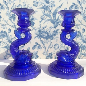 May include: A pair of cobalt blue glass candle holders shaped like a dolphin. The candle holders have a detailed design and a textured base.