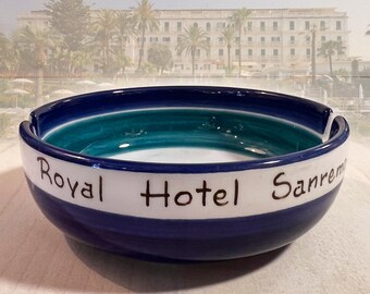 May include: A blue and white ceramic ashtray with the words "Royal Hotel Sanremo" written on it. The ashtray has a teal green stripe around the inside.