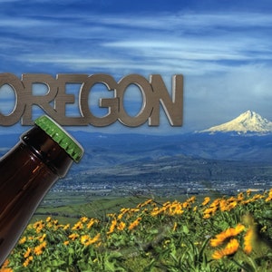 May include: A metal bottle opener with the word "OREGON" in large letters. The opener is in front of a scenic view of a mountain range with a snow-capped peak and a field of yellow wildflowers.
