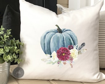 May include: A white pillow with a blue watercolor pumpkin and floral design. The pumpkin is surrounded by white and burgundy flowers and feathers.