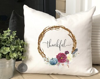 May include: White throw pillow with a wreath design featuring fall foliage, flowers, and pumpkins. The wreath has the word "thankful" written in cursive inside.