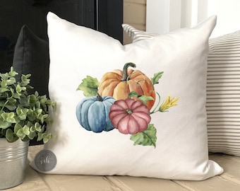 May include: A white pillow with a watercolor design of three pumpkins. The pumpkins are orange, blue, and pink. The pillow is perfect for fall decor.