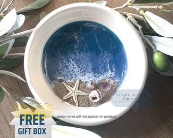 May include: A white ceramic bowl with a blue and white resin ocean scene inside. The scene includes white foamy waves, blue water, and a sandy beach with a starfish and seashells.