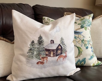 May include: A white throw pillow with a watercolor design of a cabin in the woods with deer. The cabin has a brown roof and a chimney with smoke coming out of it. The trees are green and the ground is covered in snow. The pillow is on a brown leather couch.