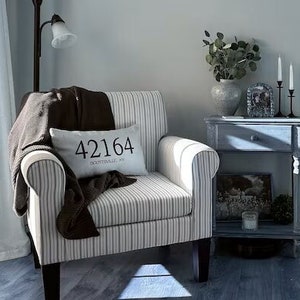 May include: A living room interior with a striped armchair, a throw, and a cushion with the text "42164". A floor lamp, a side table with ornaments, and a faux olive tree in a basket create a comfortable atmosphere.