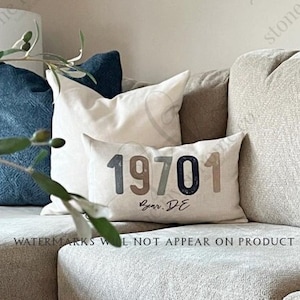 May include: A light gray sectional sofa with a throw pillow featuring the text "19701" and "Year DE". The sofa is in a living room setting with a wooden coffee table and a green plant.