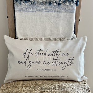 Może przedstawiać: Biała poduszka z czarnym tekstem, kt&oacute;ry brzmi "He stood with me and gave me strength 2 Timothy 4:17". Poduszka leży na plecionym krześle.